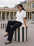 A woman in a white half-zip cotton polo and black wide trousers sits on a striped stone column in the Palais-Royal courtyard on an overcast morning.