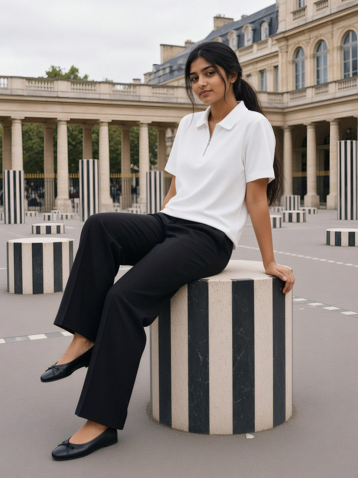 A woman in a white half-zip cotton polo and black wide trousers sits on a striped stone column in the Palais-Royal courtyard on an overcast morning.