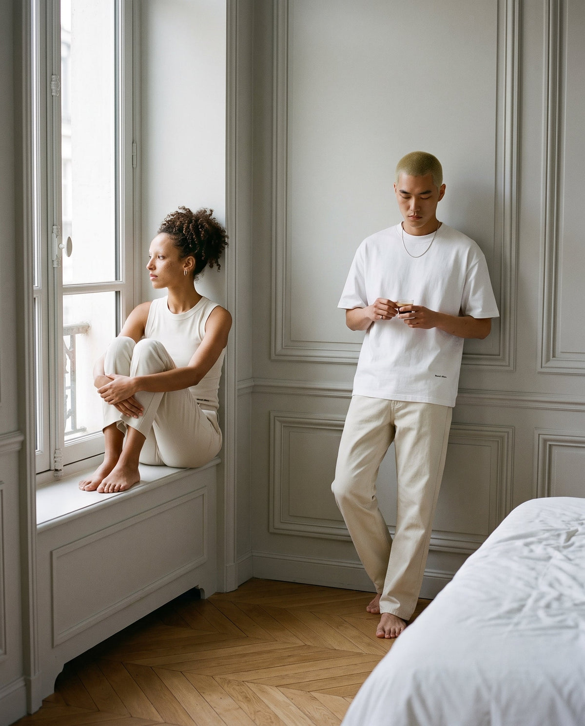 Two friends in Marcel Afters Essentials in white and bone, a ribbed tank and a heavyweight tee, in a quiet Paris apartment.