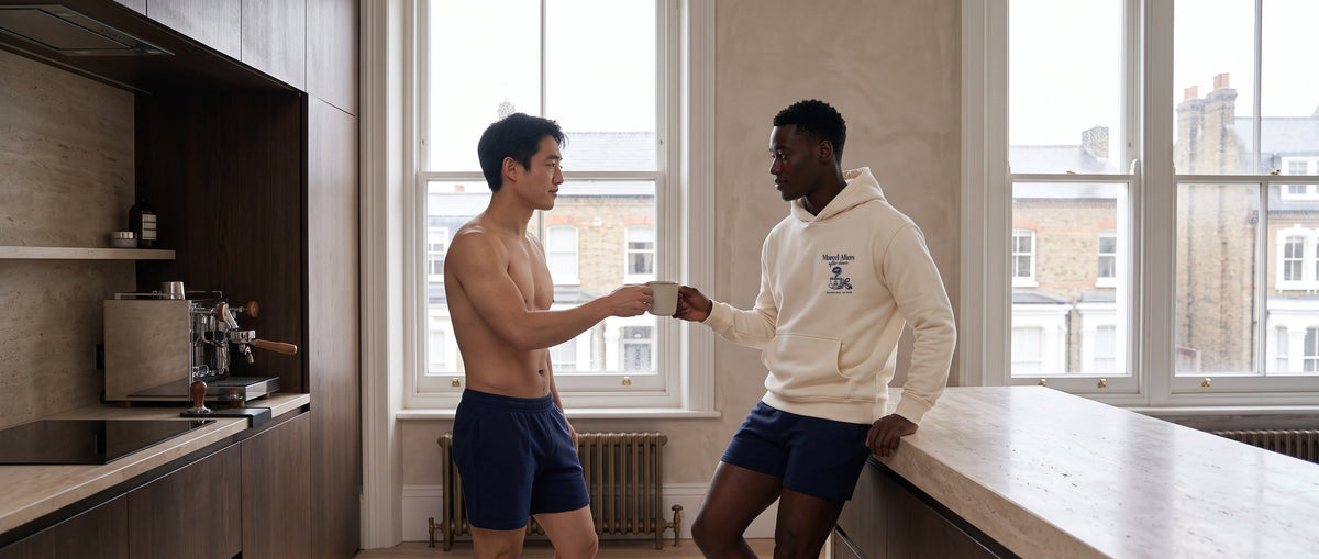Two men sharing coffee in a London kitchen, one in the cream Marcel Afters Morning After hoodie with a navy left-chest print.