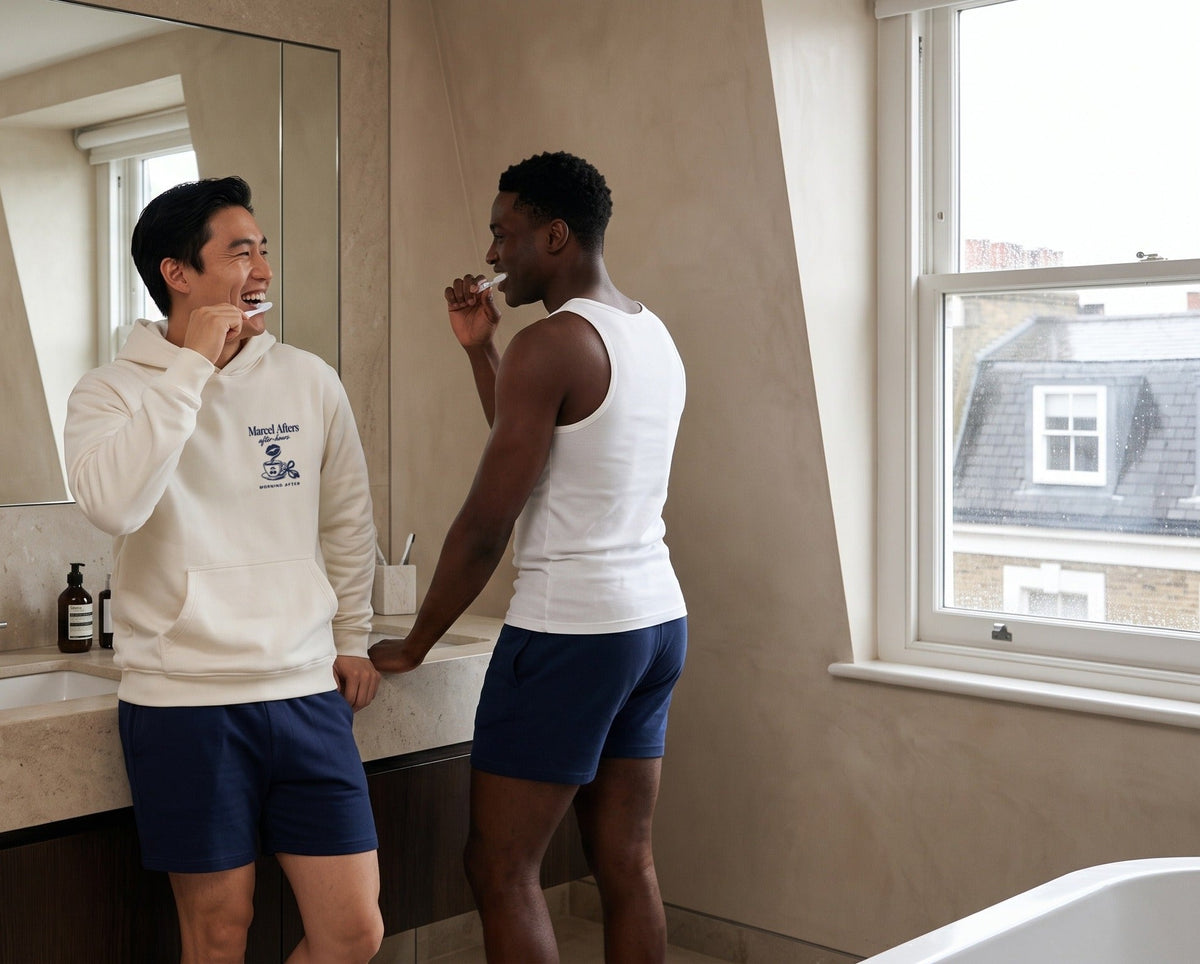 Two men brushing their teeth and laughing at a stone vanity, one in the cream Marcel Afters Morning After hoodie.