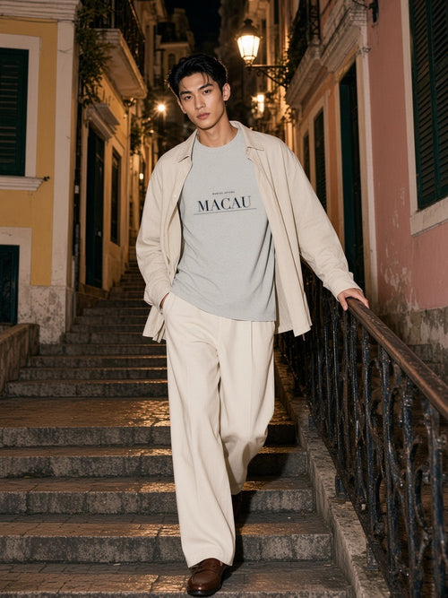 Man in the Macau tee in Oat Gray under a beige trench coming down stone steps in a Macau lane at night