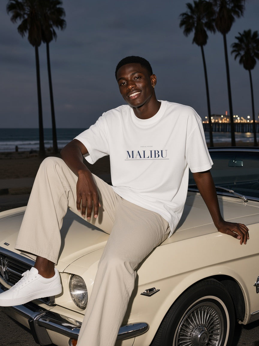 Man in the Malibu Heavyweight Drop Shoulder T-Shirt in White with the navy Malibu city plate, leaning on a vintage convertible under the palms in Malibu at night