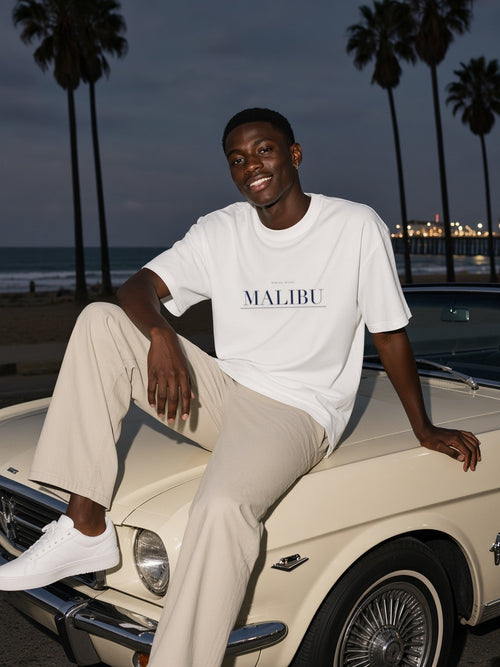 Man in the Malibu Heavyweight Drop Shoulder T-Shirt in White with the navy Malibu city plate, leaning on a vintage convertible under the palms in Malibu at night