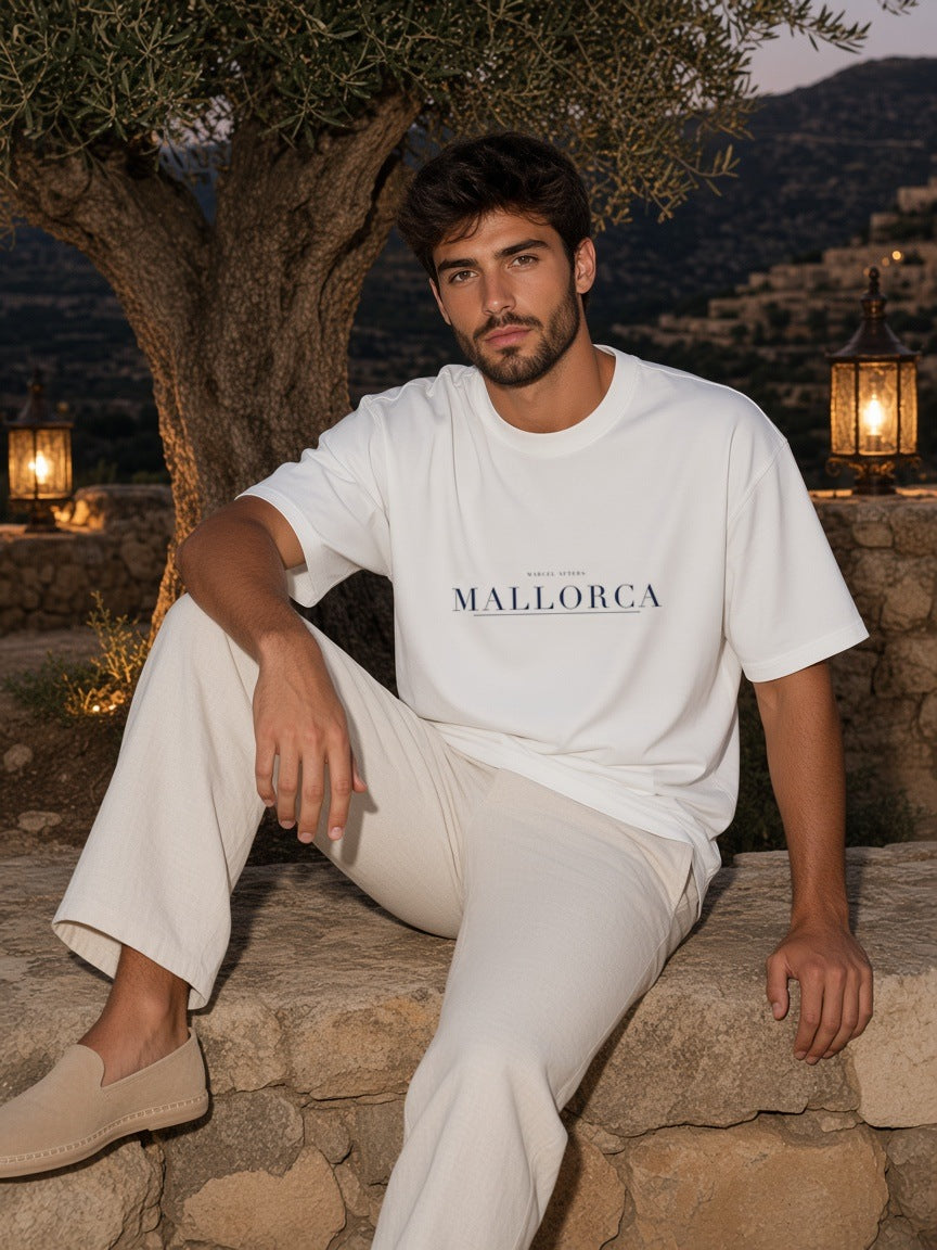 Man in the Mallorca Heavyweight Drop Shoulder T-Shirt in White with the navy Mallorca city plate, in a lantern-lit finca courtyard in Mallorca at night