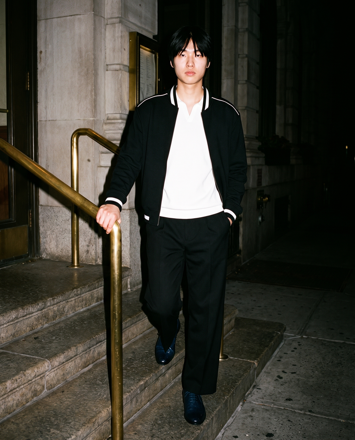 Man in Black adjustable-waist straight-leg trousers with a white knit polo and a black varsity jacket, walking down stone steps at night