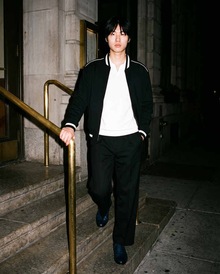 Man in Black adjustable-waist straight-leg trousers with a white knit polo and a black varsity jacket, walking down stone steps at night