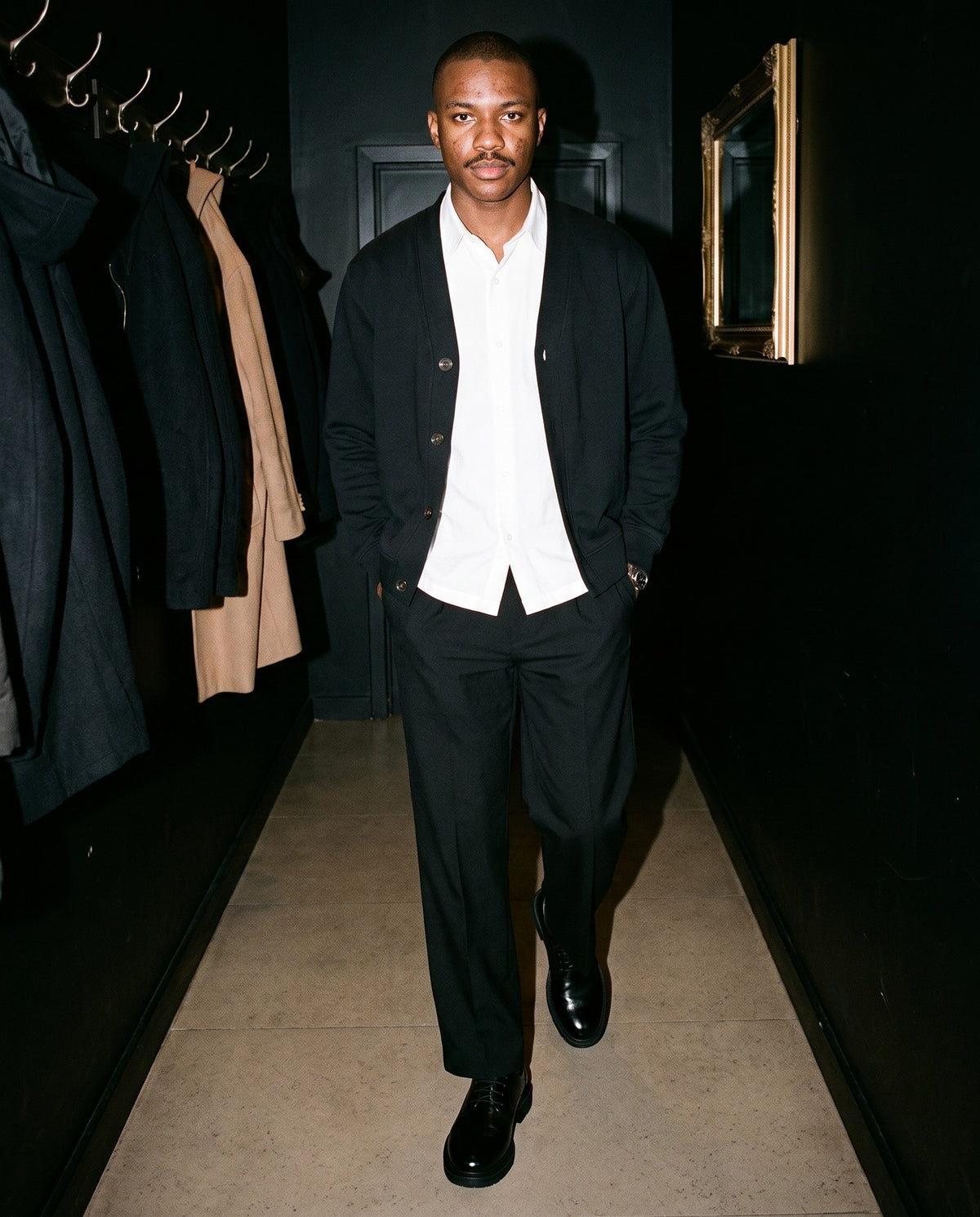 Man wearing The Night Watch Derby with the Lightweight Cotton Short Sleeve Shirt in White, Adjustable Waist Straight-Leg Pants in Black, Essential Heavyweight Sweat Cardigan in Black. Dinner at Nine, Autumn–Winter 2026 campaign