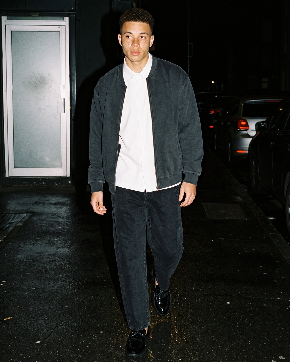 Man wearing The Ballroom Loafer with the Boxy Short Sleeve Shirt in White, Mineral Wash Barrel Leg Pants in Black, Vintage Washed Fleece Baseball Jacket in Black. After Dark, Autumn–Winter 2026 campaign