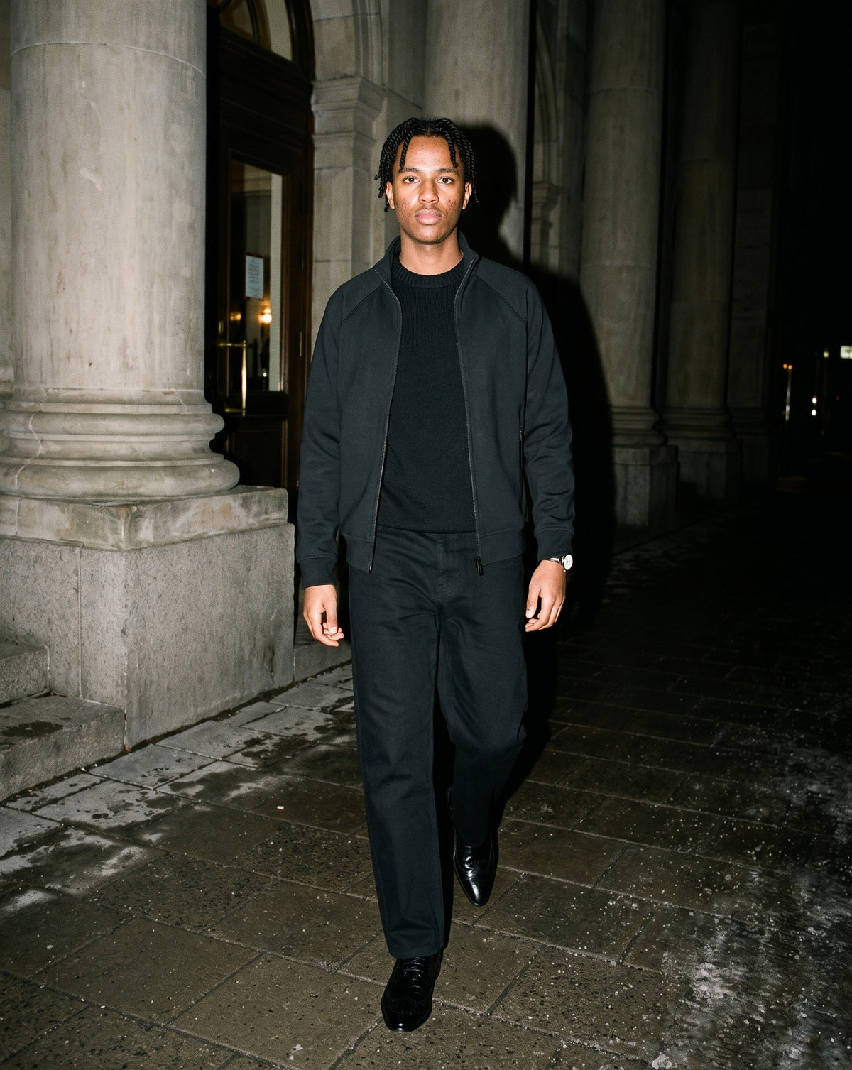 Man wearing The Head Waiter Oxford with the Knitted Round Neck Oversize Sweatshirt in Black, Heavyweight Cotton Straight-Leg Pants in Black, Zip-Pocket Stand Collar Jacket in Black. Dinner at Nine, Autumn–Winter 2026 campaign