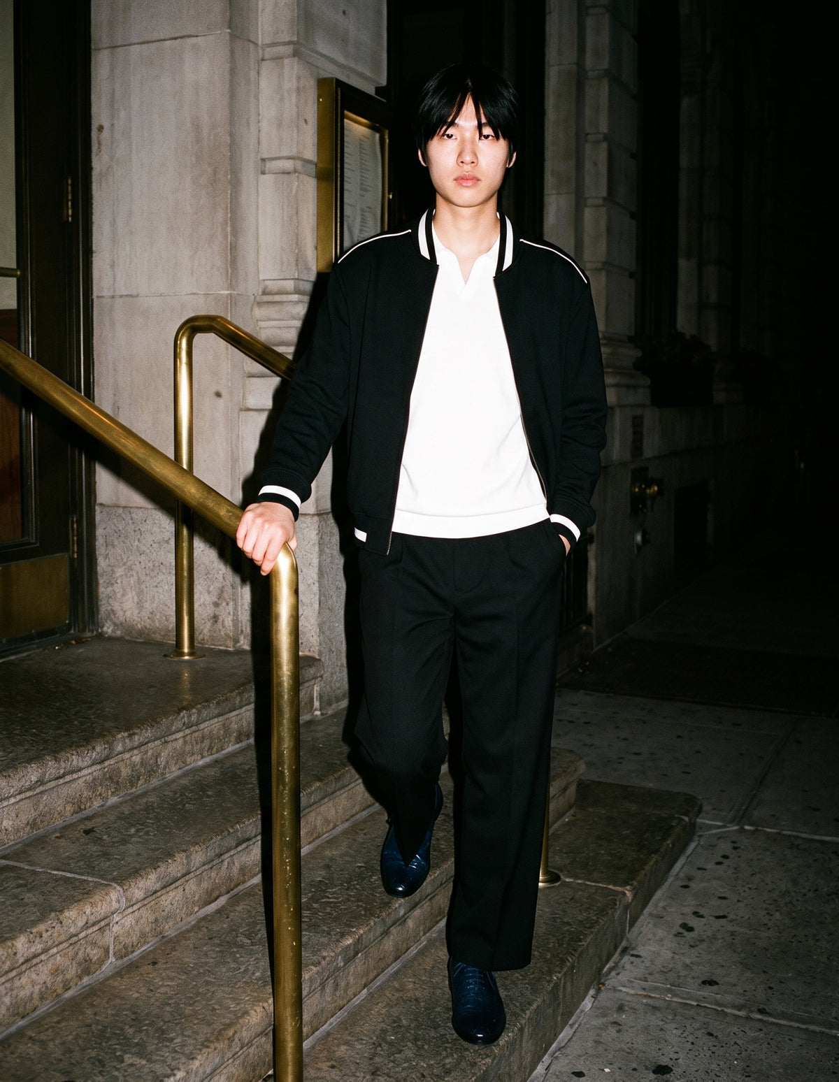 Man wearing The Blue Hour Oxford with the Heavyweight Oversized Polo in White, Adjustable Waist Straight-Leg Trousers in Black, Contrast Piping Zip Bomber Jacket in Black. Dinner at Nine, Autumn–Winter 2026 campaign