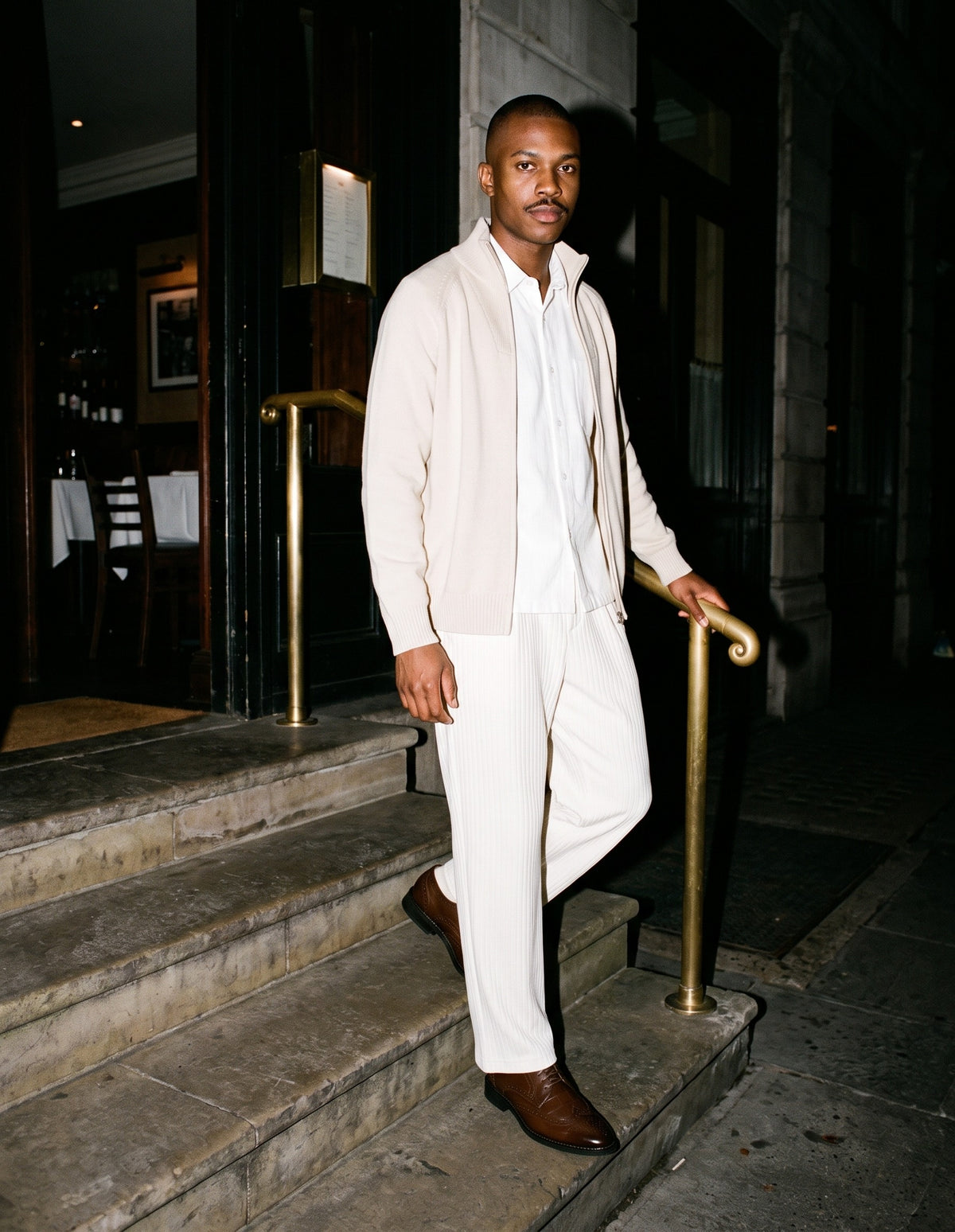 Man wearing The Library Bar Brogue with the Boxy Heavyweight Short Sleeve Shirt in White, Striped Pleated Pants in White, Quarter-Zip Pullover Sweatshirt in Beige. Dinner at Nine, Autumn–Winter 2026 campaign