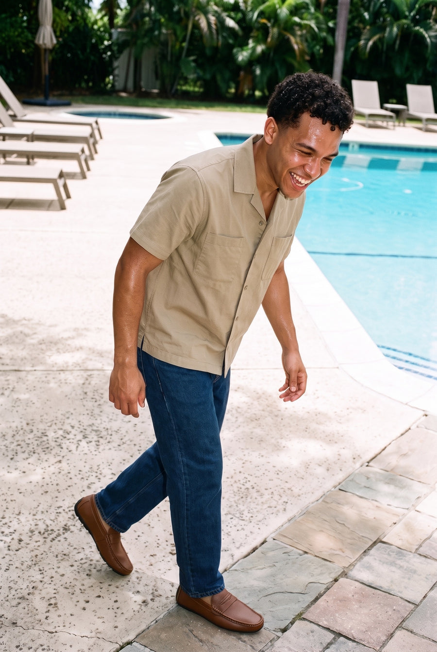 Man laughing beside a swimming pool in a khaki camp-collar shirt and dark blue jeans wearing The Brown Loafer in brown leather, full length