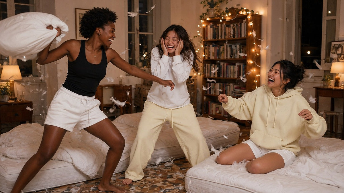 Three women in ribbed tanks, sweatshorts and waffle tops mid pillow fight on floor mattresses.