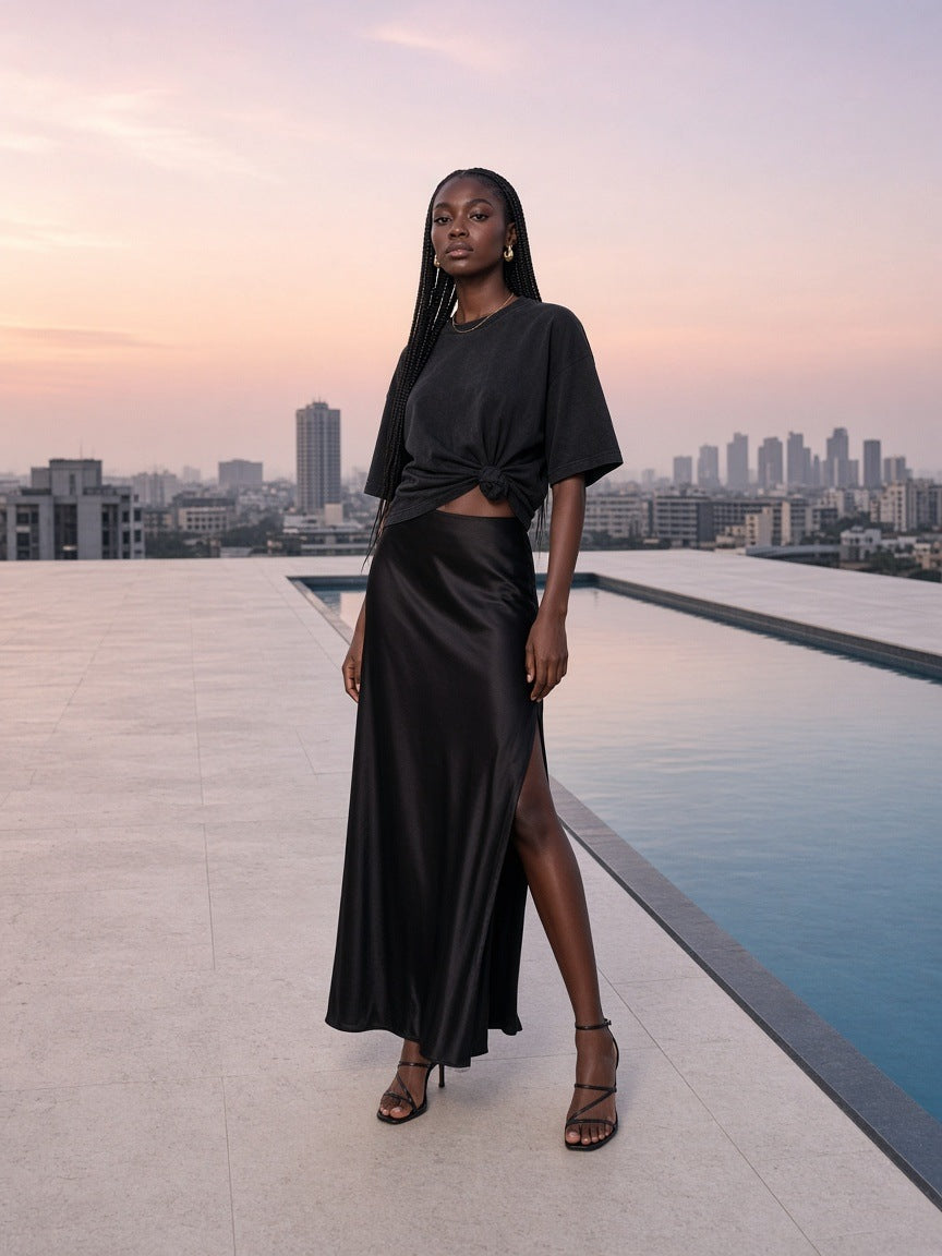 Marcel Afters Classic Loose Boxy Tee in Black, worn by a woman, standing by a rooftop pool at sunset in a long satin skirt