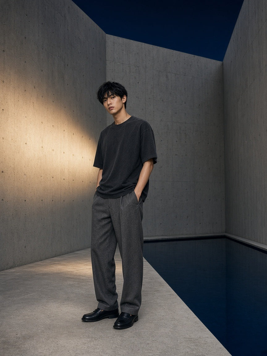 Marcel Afters Classic Loose Boxy Tee in Black, worn by a man, standing in a concrete corner beside dark water in grey trousers