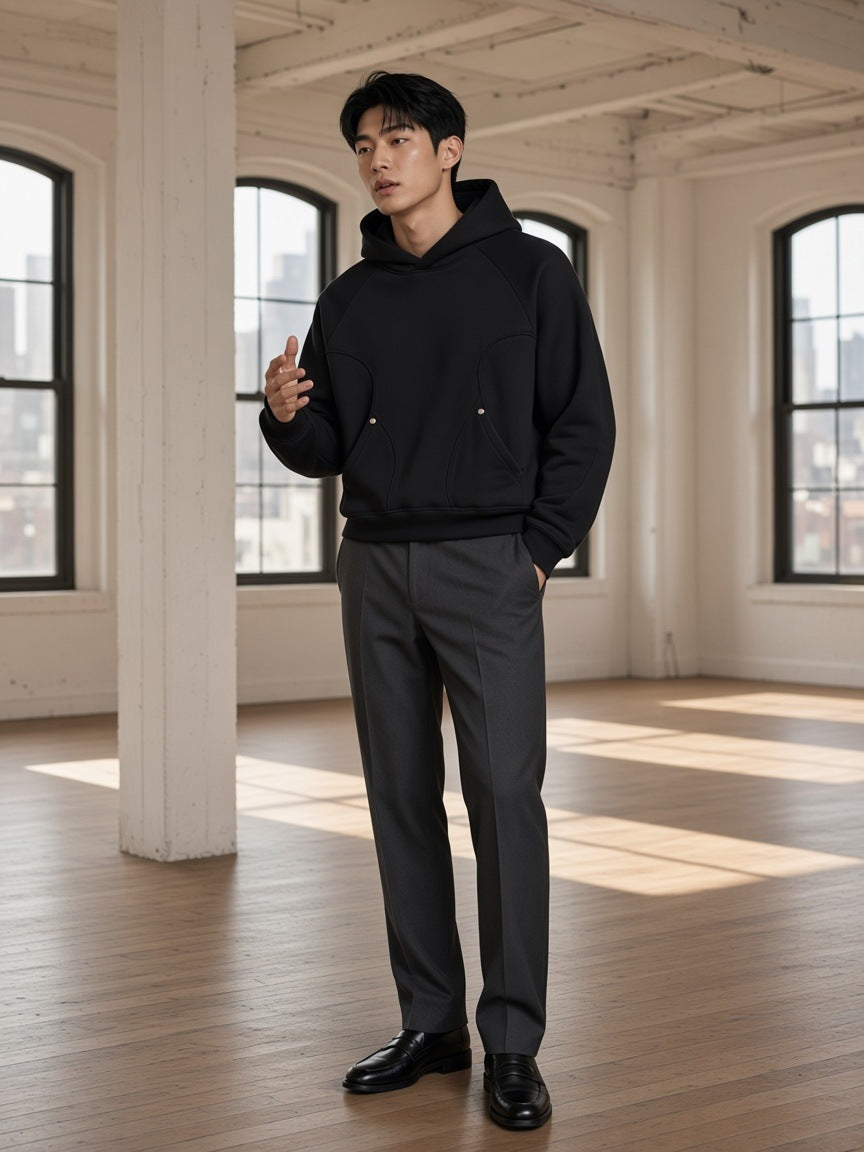 Marcel Afters Curved Panel Raglan Hoodie in Black, worn by a man, standing in a white loft with tall windows in grey trousers