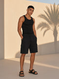 Marcel Afters Drawstring Cargo Shorts in Black, worn by a man, standing barefoot against a sunlit wall with palm shadows in a black tank
