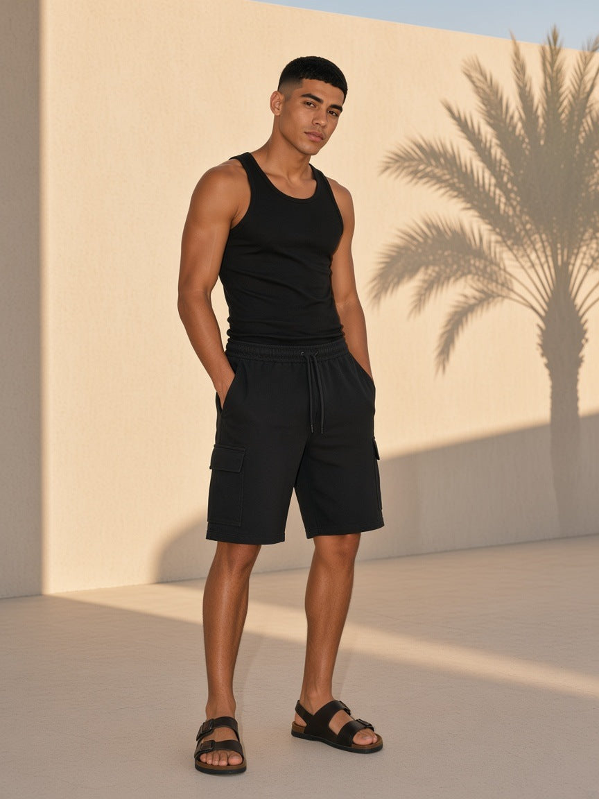 Marcel Afters Drawstring Cargo Shorts in Black, worn by a man, standing barefoot against a sunlit wall with palm shadows in a black tank