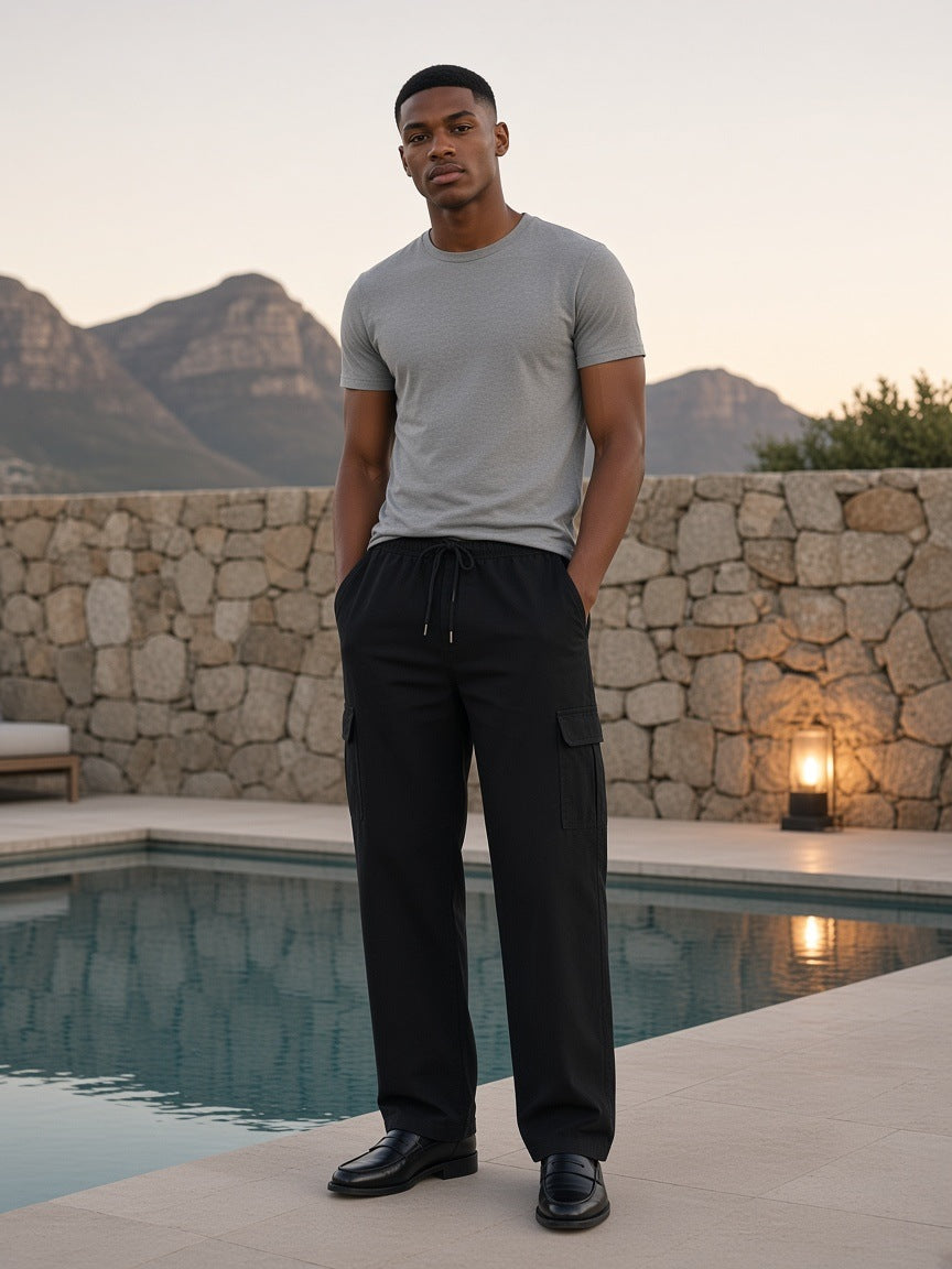 Marcel Afters Drawstring Waist Straight-Leg Cargo Pants in Black, worn by a man, standing by a rocky-view pool at dusk in a grey tee