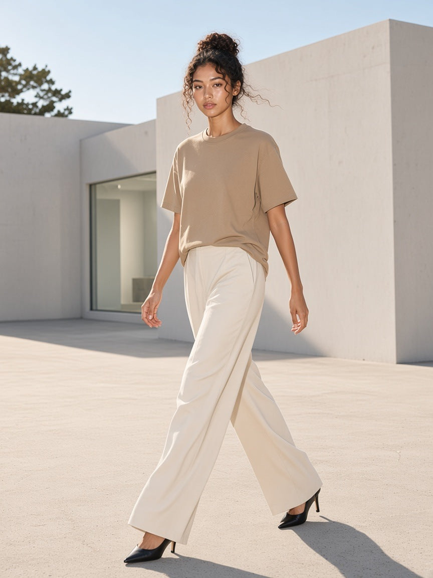 Marcel Afters Enzyme Washed T-Shirt in Camel, worn by a woman, walking a sunlit concrete plaza in wide cream trousers and heels