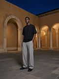 Marcel Afters Essential Cotton T-Shirt in Black, worn by a man, standing in a sandstone arcade at night in grey trousers