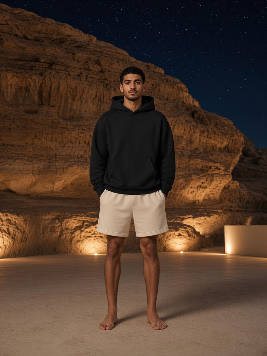 Marcel Afters Essential Heavyweight Boxy Fleece Hoodie in Black, worn by a man, standing barefoot in cream shorts before a lit cave wall at night