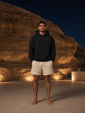 Marcel Afters Essential Heavyweight Boxy Fleece Hoodie in Black, worn by a man, standing barefoot in cream shorts before a lit cave wall at night