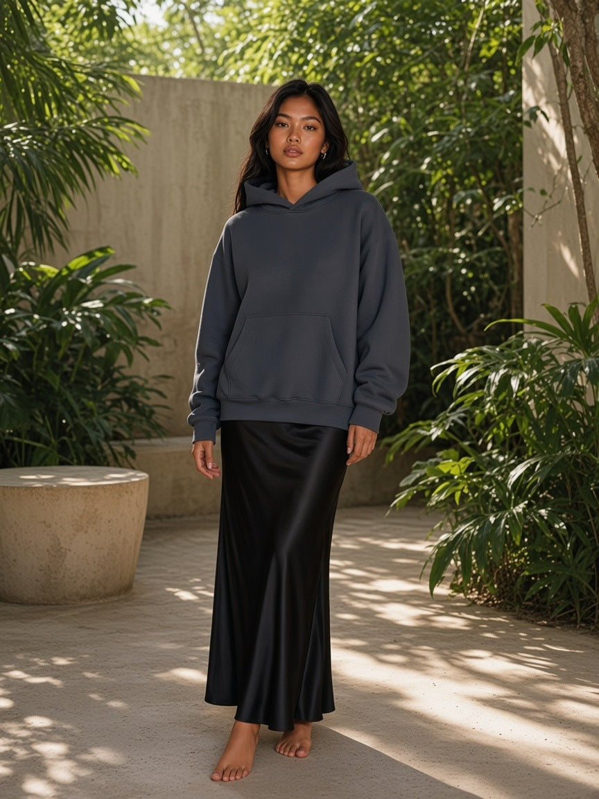 Marcel Afters Essential Heavyweight Boxy Fleece Hoodie in Gray Blue, worn by a woman, standing in a sunlit jungle courtyard in a long black satin skirt