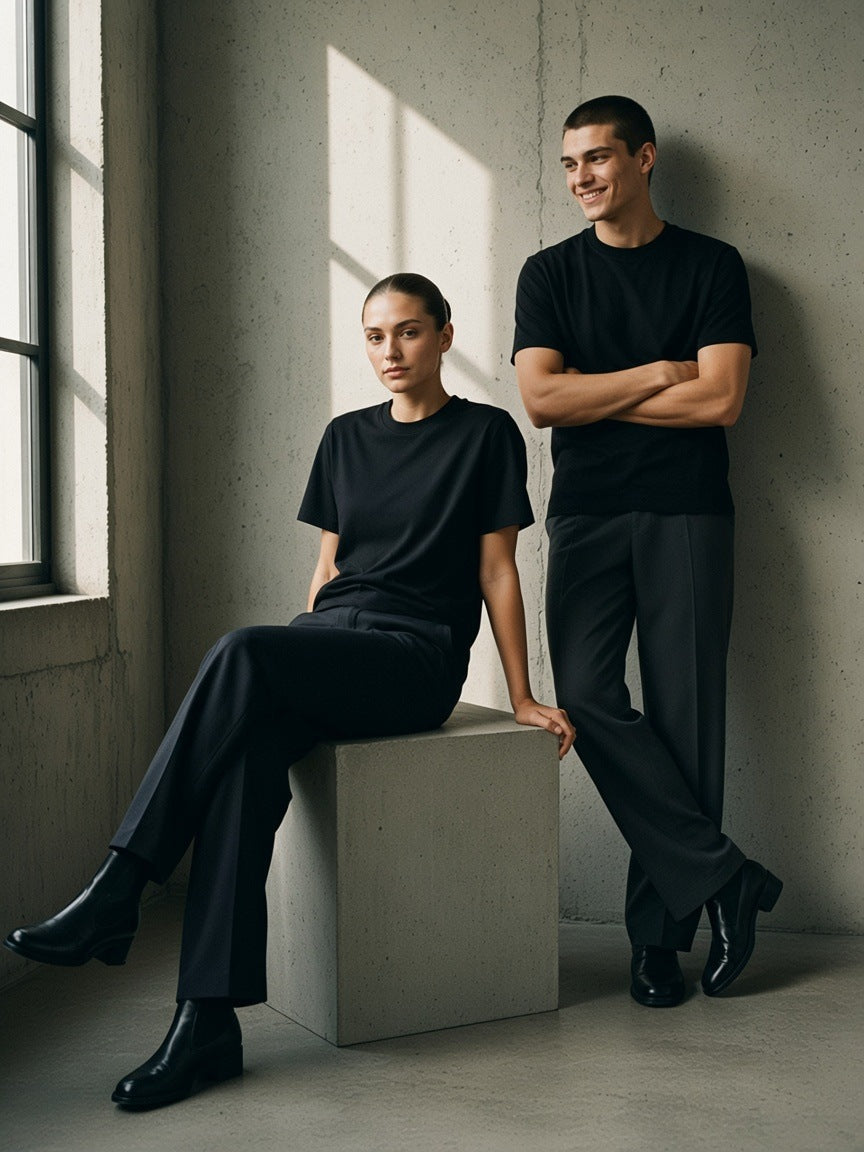 Two people in the Essential Heavyweight Cotton T-Shirt in Black, one seated on a concrete plinth, one standing, in a sunlit concrete studio in the morning