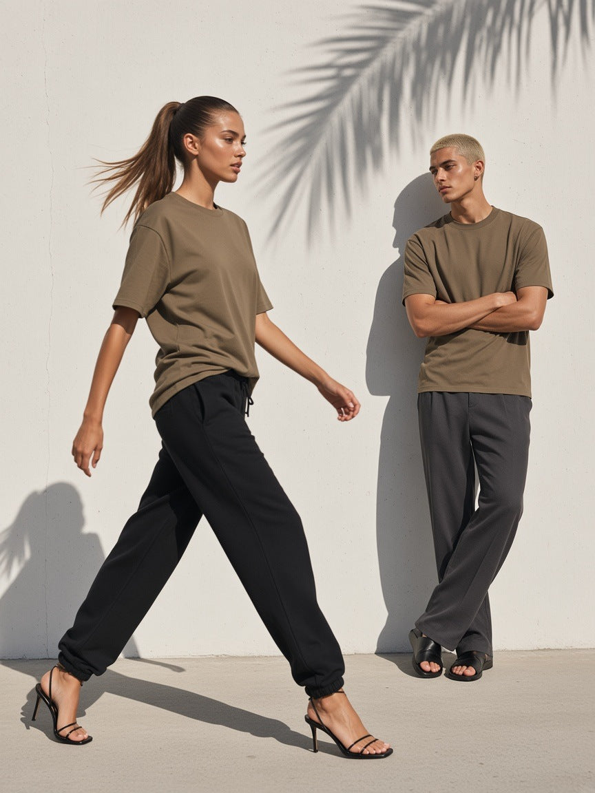 Two people in the Essential Heavyweight Cotton T-Shirt in Moss Green, a woman striding in heels and black sweatpants past a white concrete wall with palm shadow, a man leaning behind