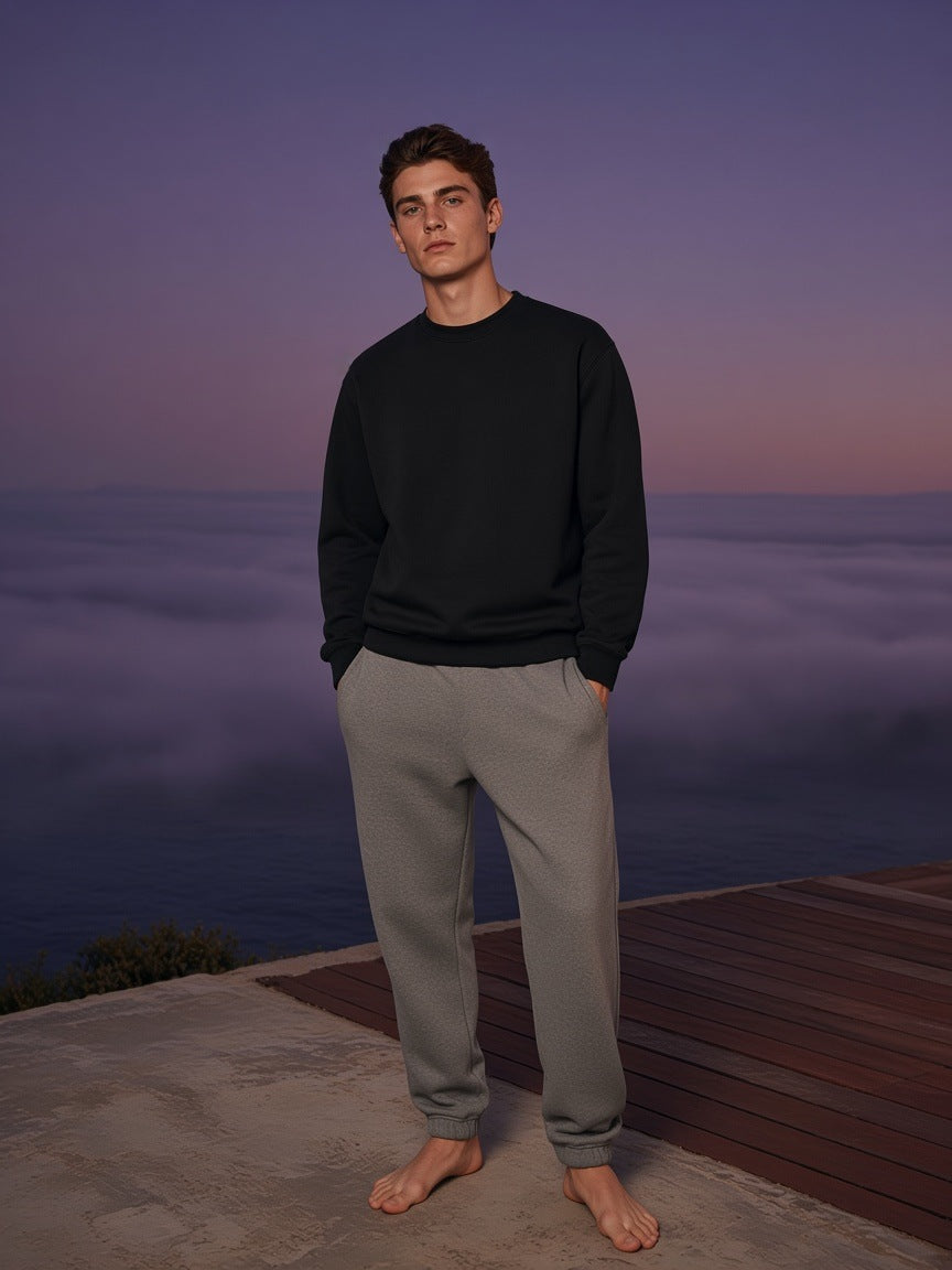 Marcel Afters Essential Heavyweight Crewneck Sweatshirt, 400 gsm in Black, worn by a man, standing barefoot on a cloud-level terrace at dusk in grey sweatpants