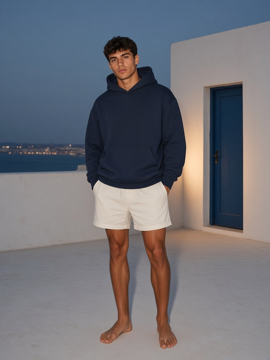 Marcel Afters Essential Heavyweight Hoodie, 400 gsm in Navy Blue, worn by a man, standing barefoot on a white terrace at dusk in white shorts