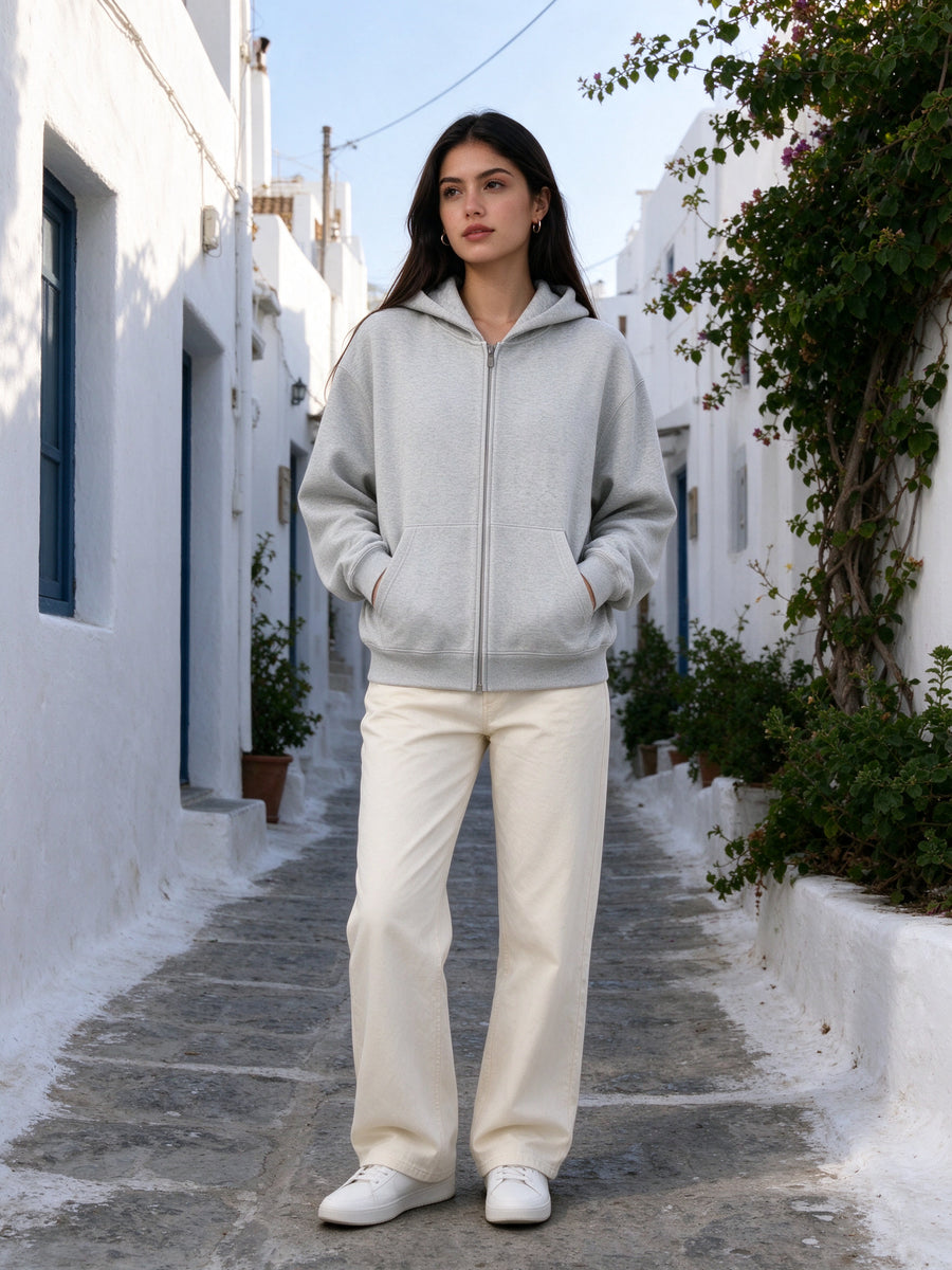 Marcel Afters Essential Heavyweight Pocket Hoodie in Flower Gray, worn by a woman, in a sunlit whitewashed Greek island lane with her hands in the pocket of a heather grey pocket hoodie