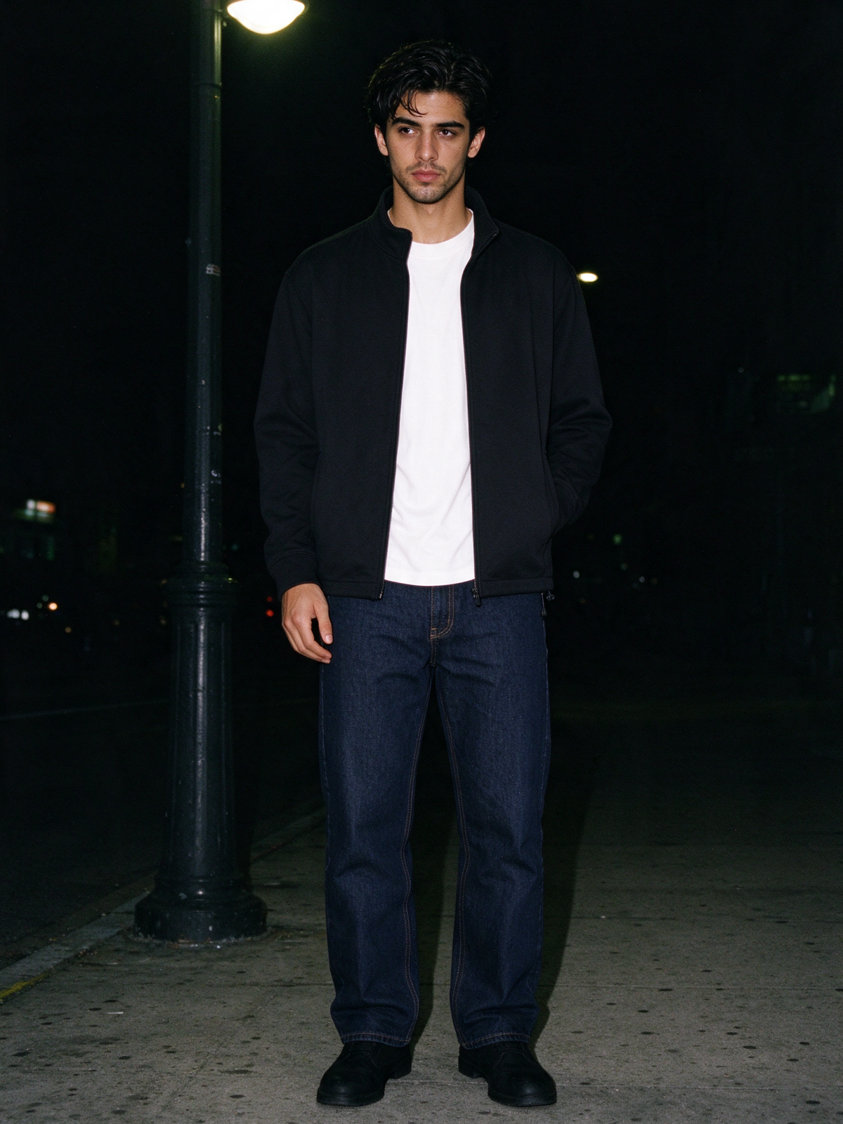 Man in Black heavyweight stand collar jacket over a white T-shirt and dark jeans, under a street lamp at night