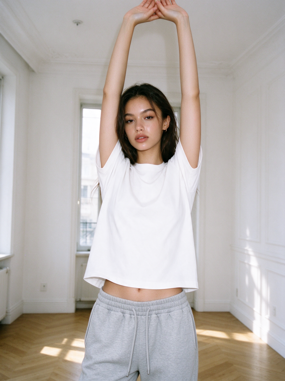 Woman in Flower Gray essential jogged sweatpants and a white T-shirt, arms raised, in a bright apartment