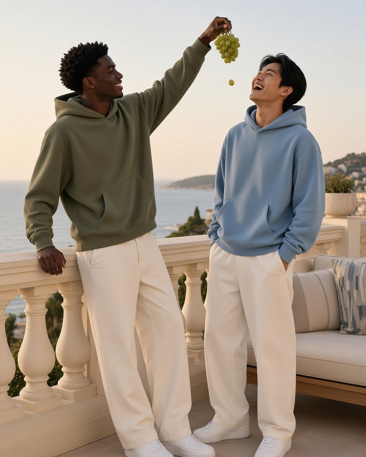 Two men in the Essential Oversized Hoodie in Army Green and Haze Blue, one dropping a grape into the other's mouth on a Riviera terrace
