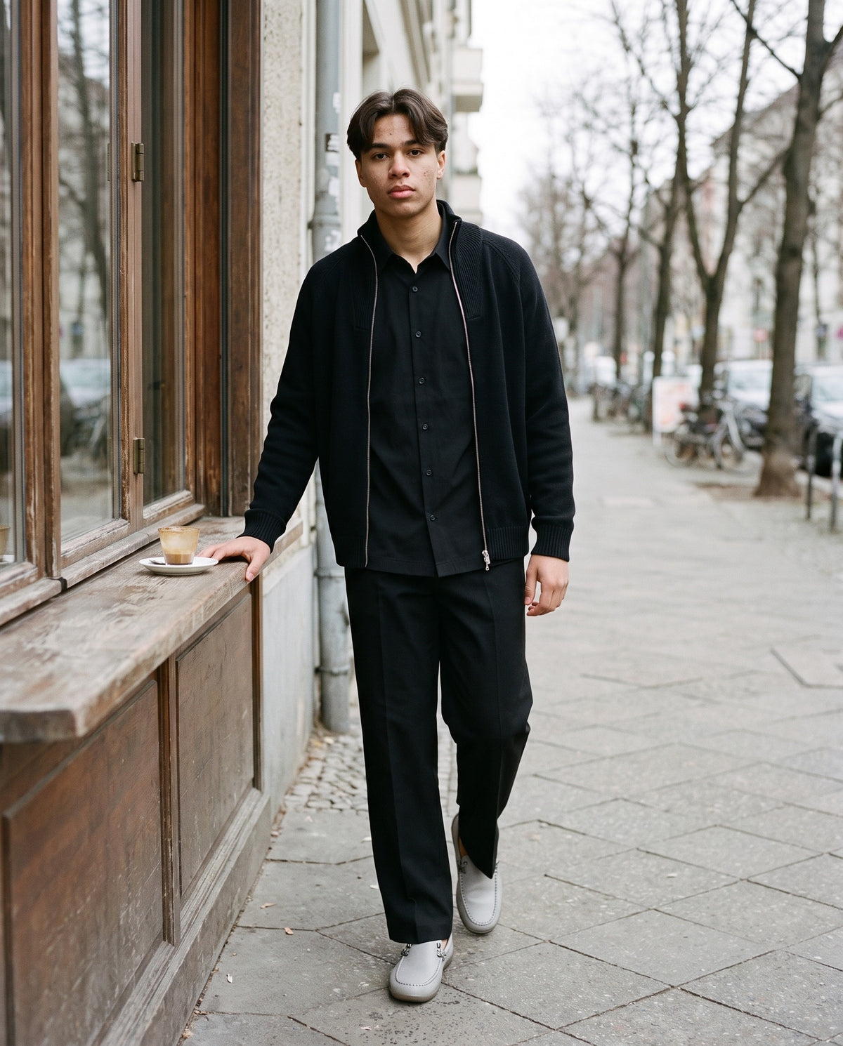 Man wearing The Sunday Papers Loafer with the Boxy Short Sleeve Shirt in Black, Adjustable Waist Straight-Leg Trousers in Black, Quarter-Zip Pullover Sweatshirt in Black. The Long Lunch, Autumn–Winter 2026 campaign