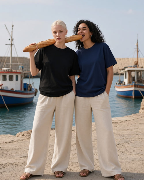 Two women in the Heavyweight Earth Tone T-Shirt in Black and Navy, one biting the end of a baguette on a Mediterranean harbour jetty