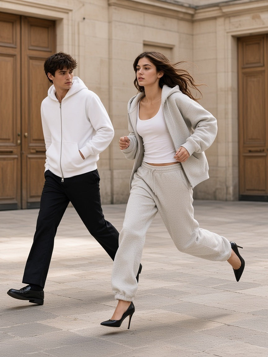 Two people in the Heavyweight Full Zip Hoodie in Oat Gray and White, a woman running in heels and oat sweatpants across a Paris limestone courtyard, a man walking behind