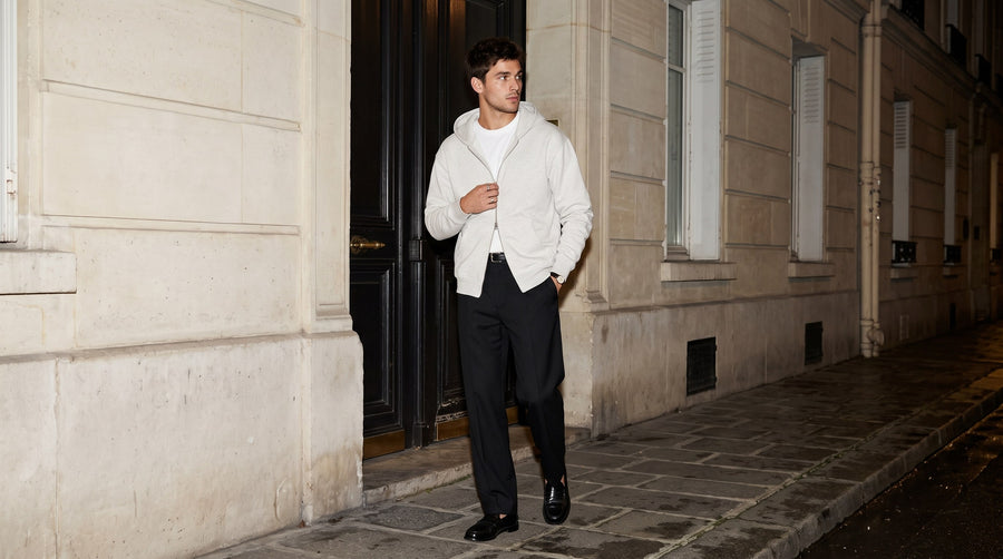 Man in the Marcel Afters Heavyweight Full Zip Hoodie in Oat Gray with black trousers and loafers stepping out of a Paris doorway at night