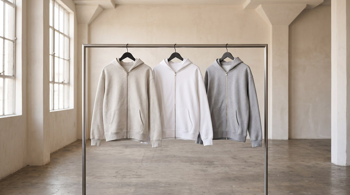 Heavyweight Full Zip Hoodie in Oat Gray, White and Flower Gray hanging on a rail in a bright stone-walled studio