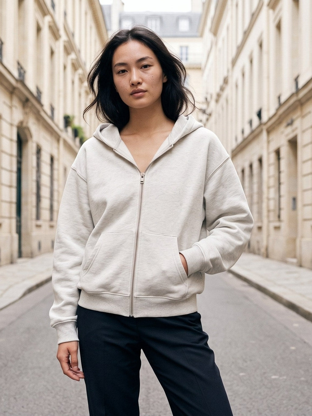 Woman in the Heavyweight Full Zip Hoodie in Oat Gray with a hand in the pocket on a Paris street