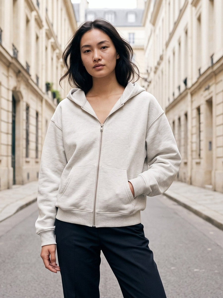 Woman in the Heavyweight Full Zip Hoodie in Oat Gray with a hand in the pocket on a Paris street