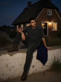 Man in the Heavyweight Snow Washed T-Shirt in Batik Black seated on a garden wall by a thatched house on Sylt at night