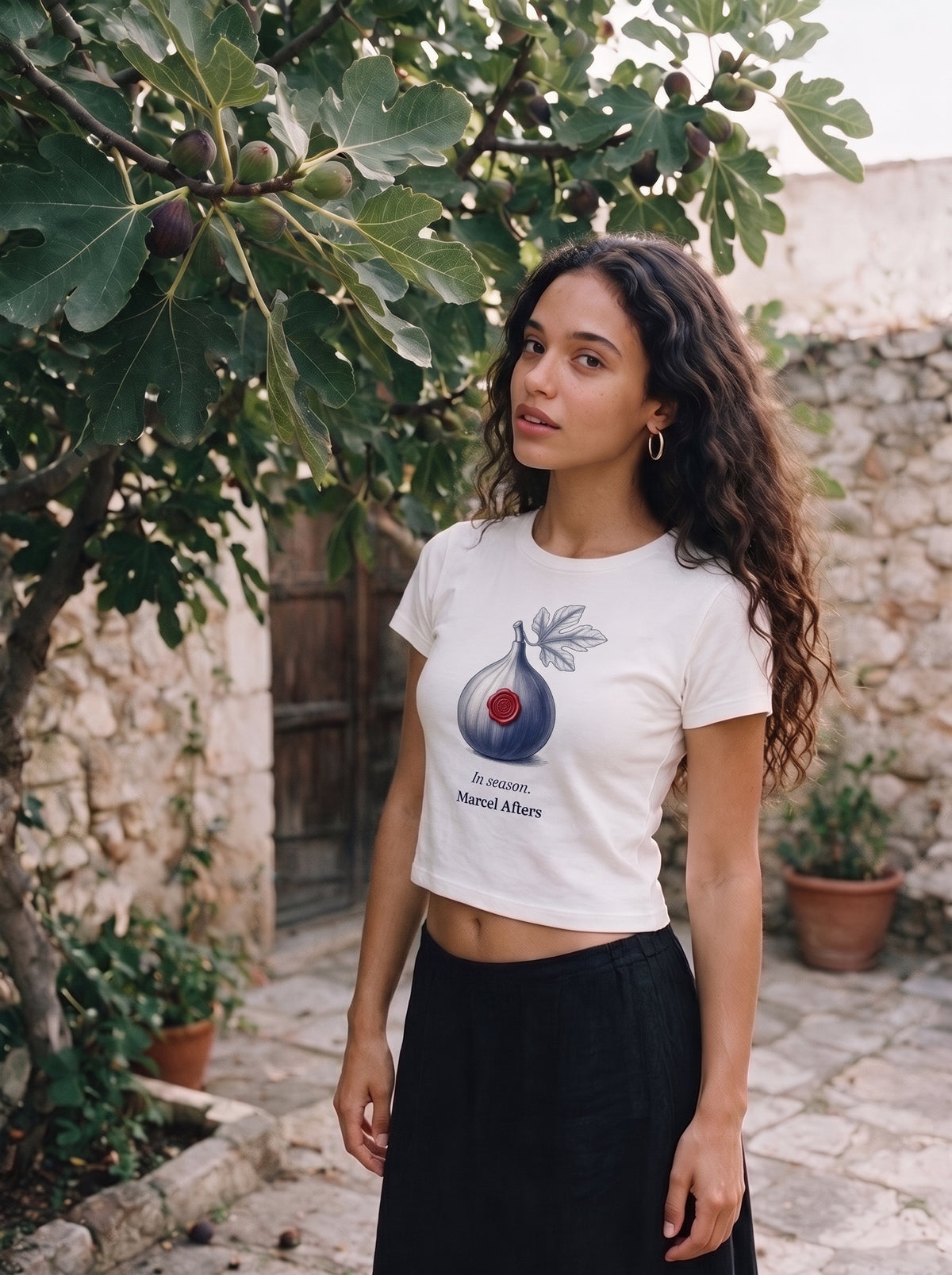 A woman standing by a fig tree in a walled courtyard in the In Season Women's Tencel Baby T-Shirt in White, fig engraving sealed with red wax on the chest, Marcel Afters