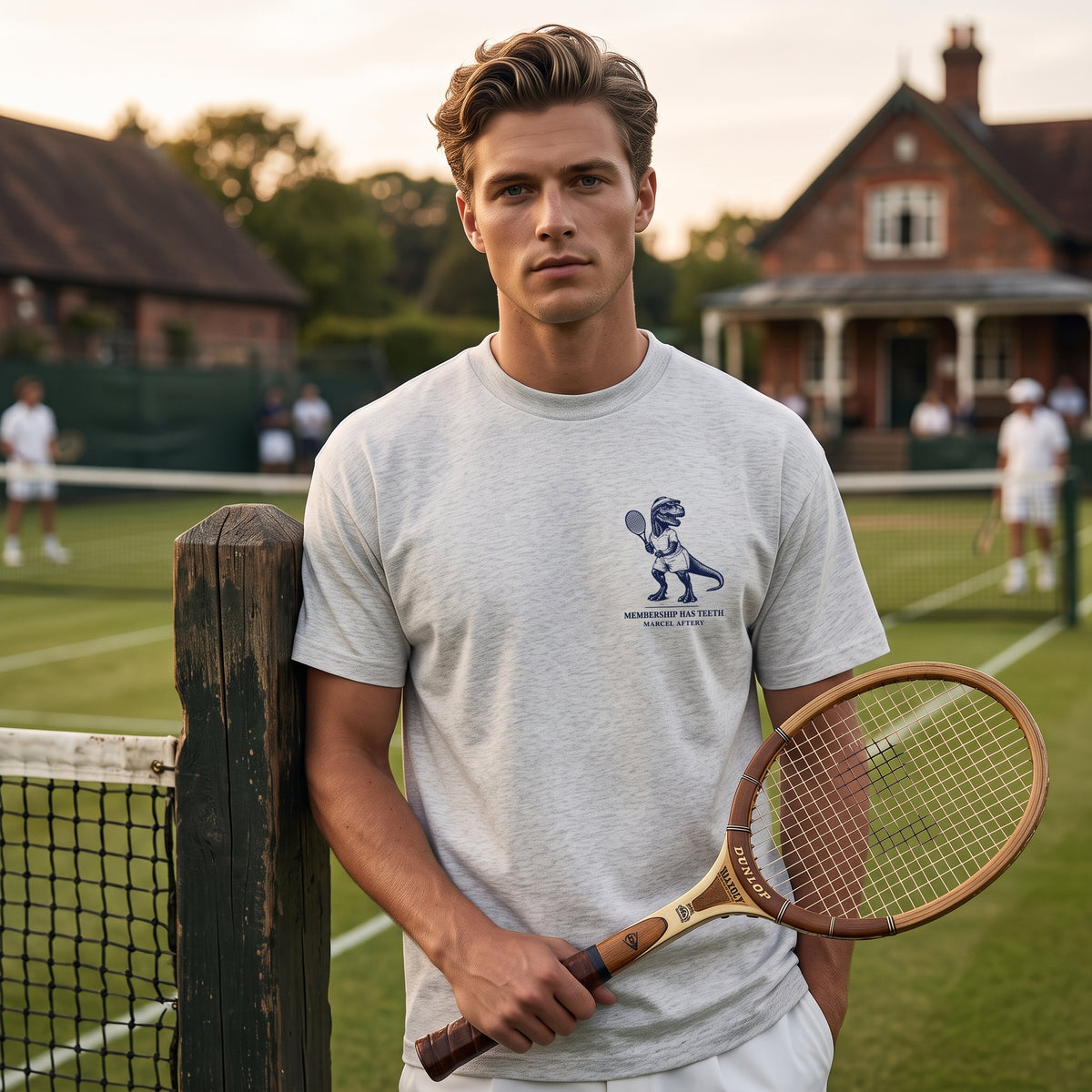 Man in the Membership Has Teeth heavyweight tee in oat gray, holding a wooden racket on a grass tennis court. Marcel Afters.