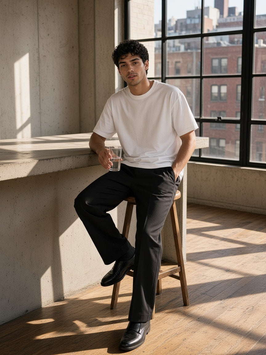 Marcel Afters Men's Oversized T-Shirt in White, worn by a man, seated on a stool by a loft window in the afternoon light