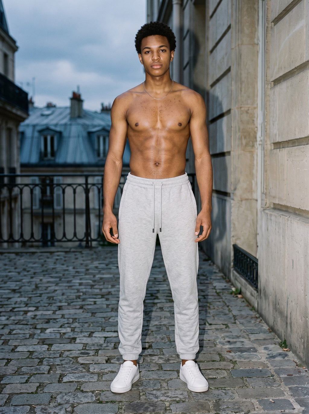 Shirtless man in the Men's Heavyweight Fleece Joggers in Flower Gray and white sneakers on Paris cobbles at dusk, lit by direct flash