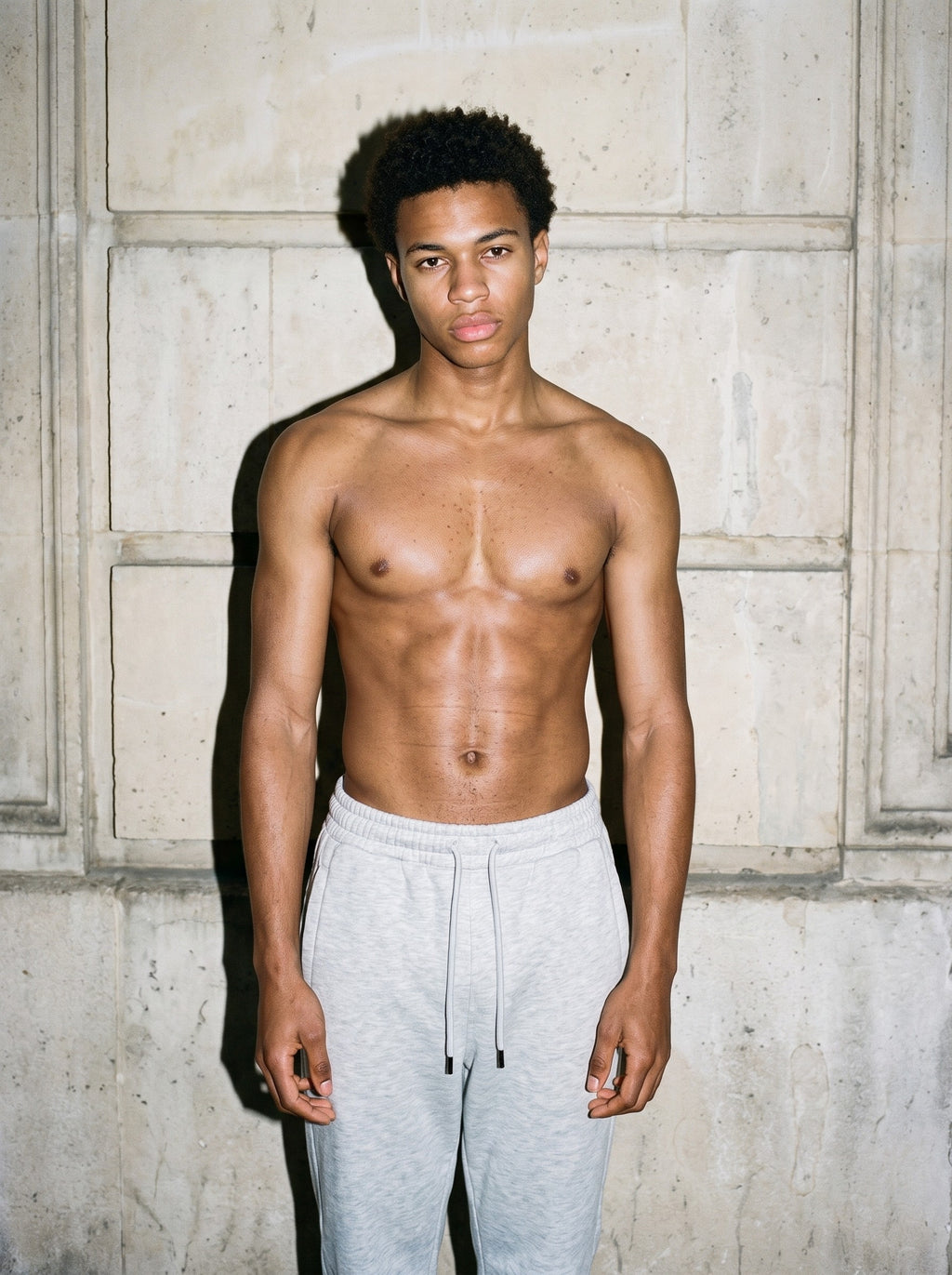 Shirtless man in the Men's Heavyweight Fleece Joggers in Flower Gray against a limestone wall, lit by direct flash